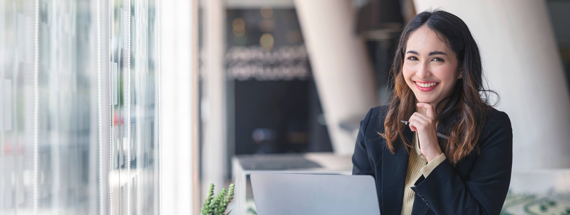 Younger business woman sitting at computer with hand by chin smiling